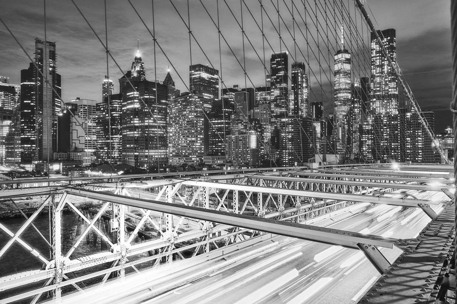 Black and white cityscape of Manhattan skyline at night with illuminated buildings viewed from Brooklyn Bridge.