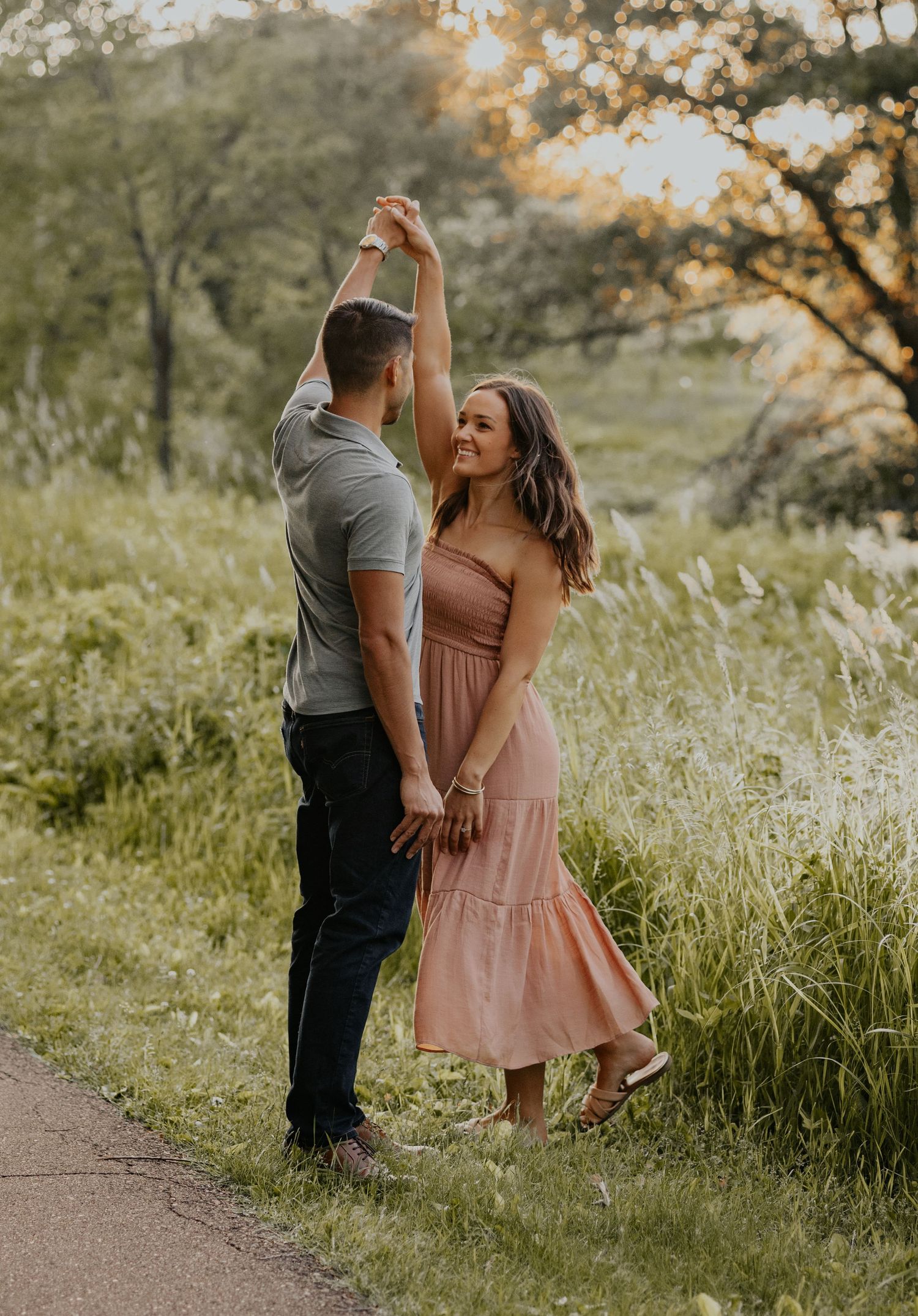 A romantic couple dances together on a path surrounded by tall grass at sunset.