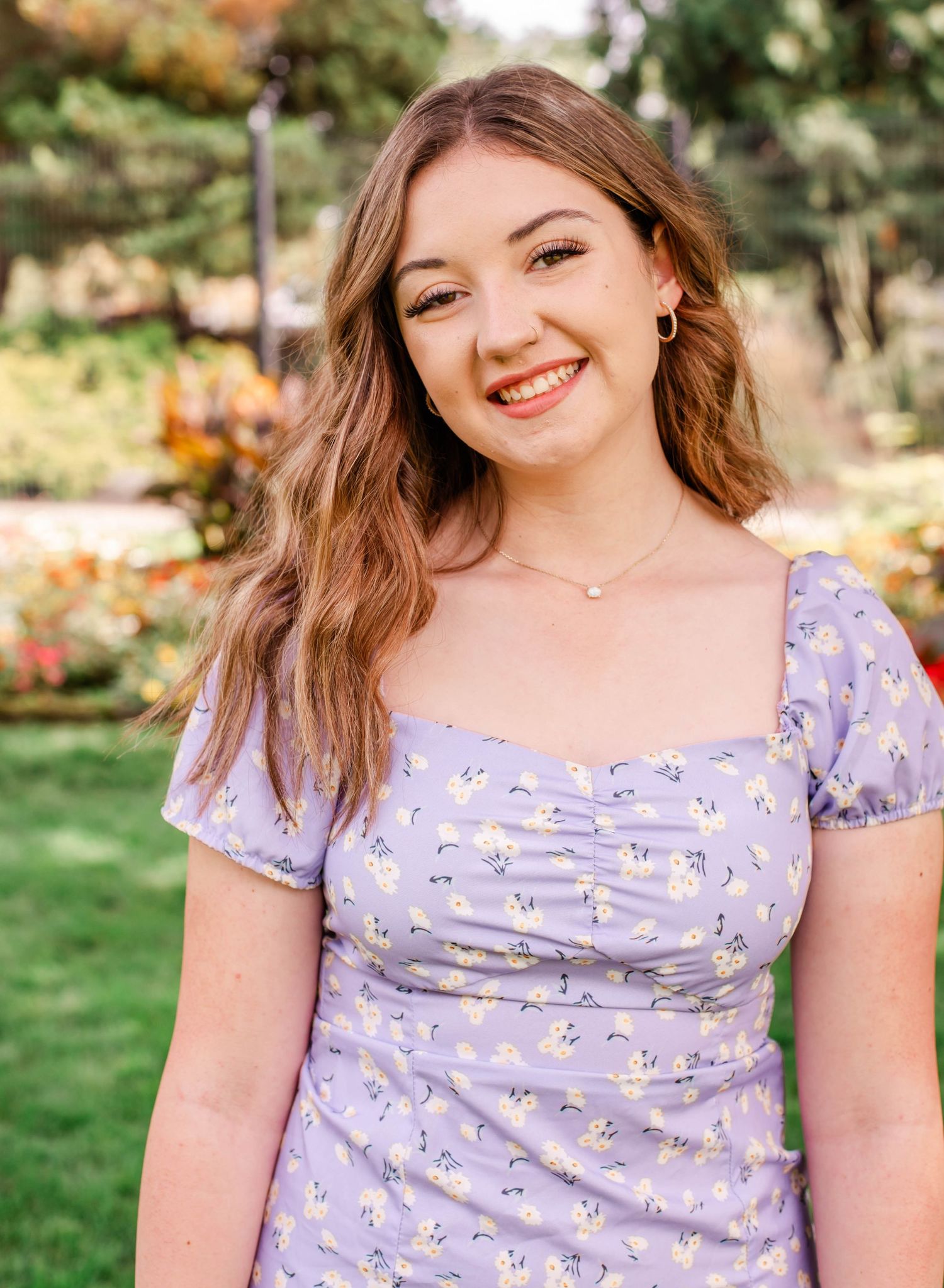 Young woman smiling in lavender floral dress outdoors in a garden setting with colorful blooms.