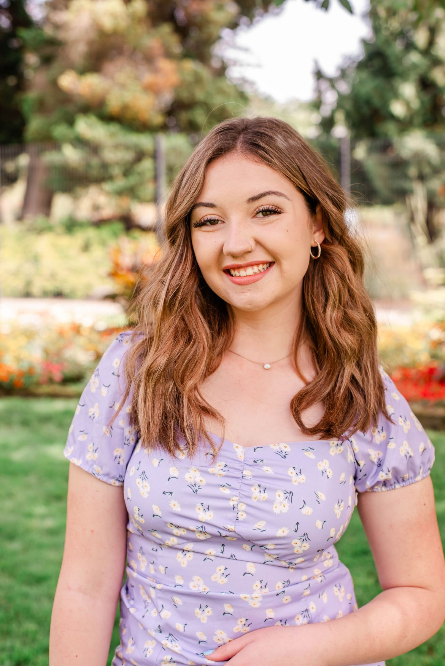 Young woman smiling outdoors in lavender floral dress with colorful garden background on sunny day.