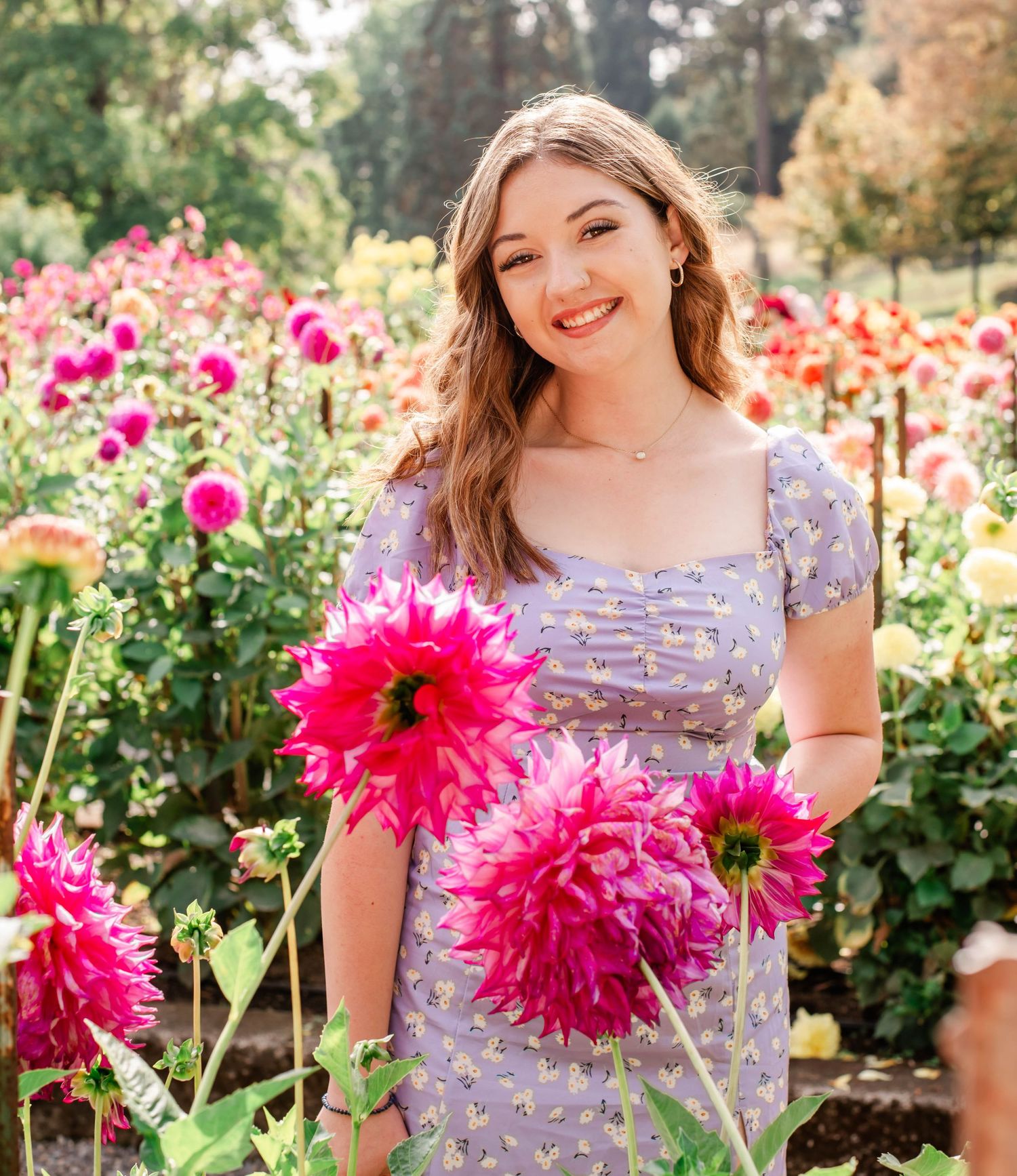 Young woman in floral dress smiling among vibrant pink dahlia flowers in a lush garden.