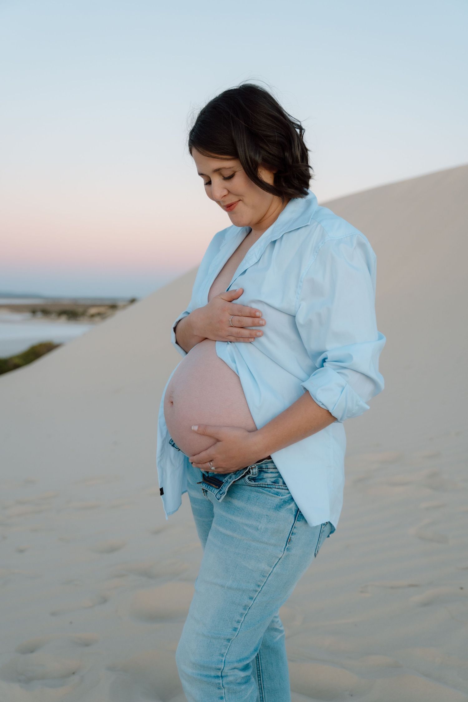 Maternity photoshoot on sandy beach at sunset showing person in light blue shirt and jeans cradling baby bump.