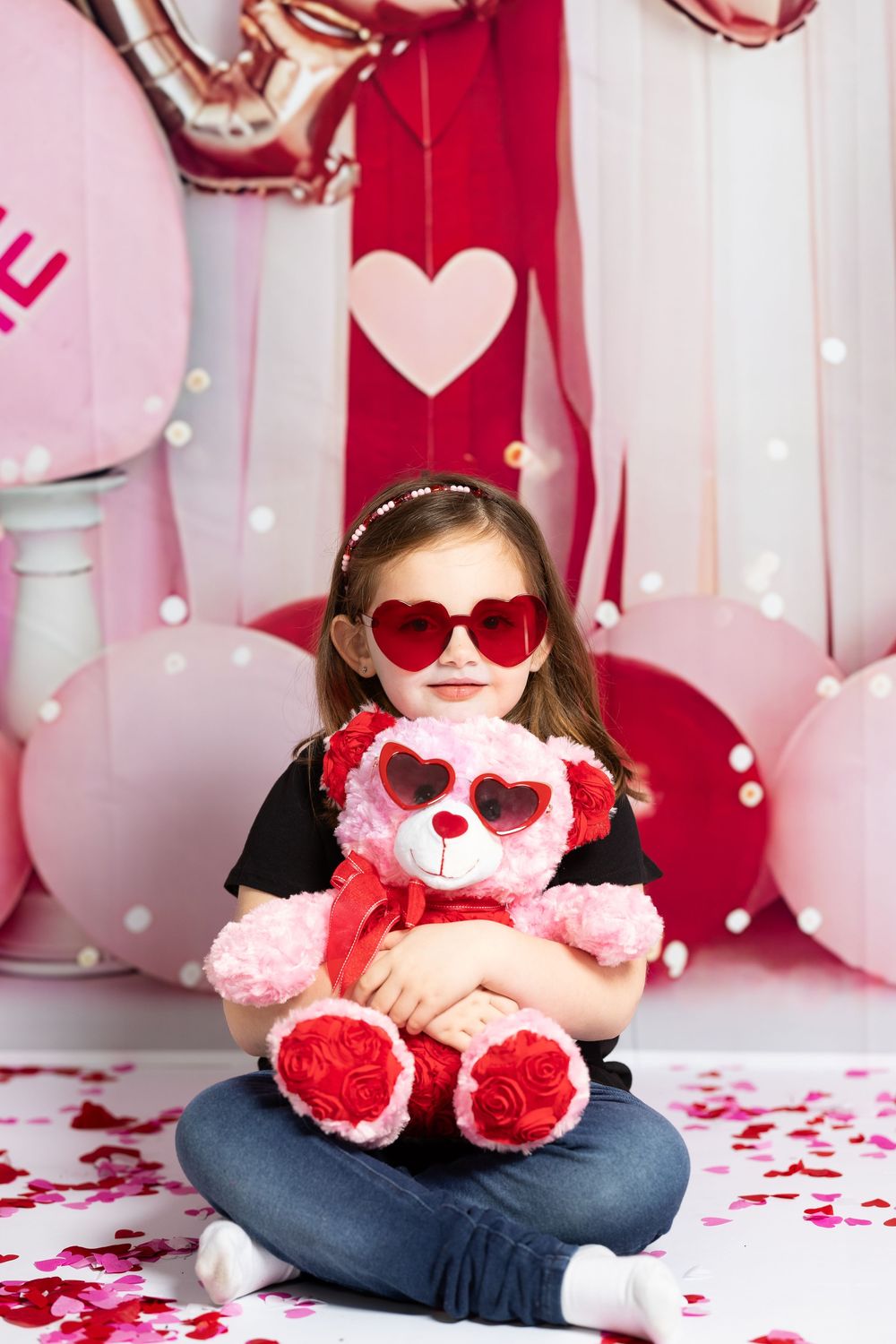 Young child wearing heart sunglasses and holding a pink teddy bear against Valentine's Day themed backdrop.