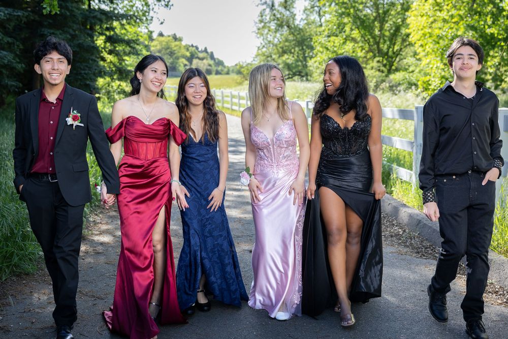 A group of young people dressed in formal attire pose together on a path during prom or homecoming event.