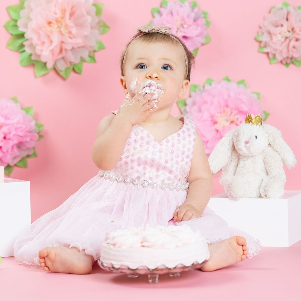 Young child in white tulle dress sitting with stuffed bunny toy against pink background with floral decorations.