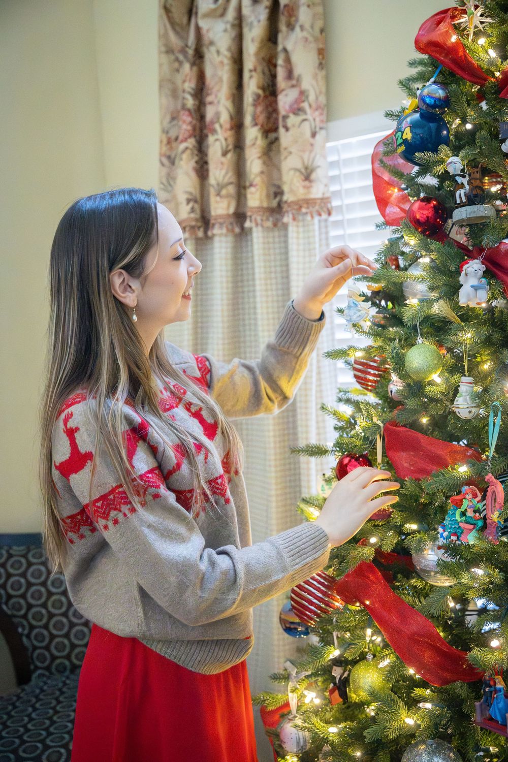 A person in a gray sweater and red skirt decorates a lit Christmas tree with ornaments and ribbons.