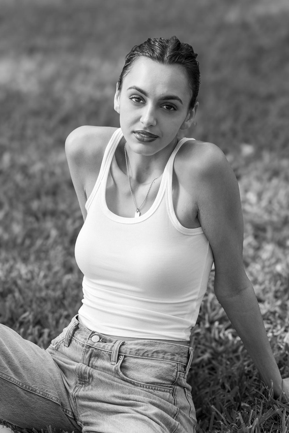 Black and white portrait of a young woman in white tank top and jeans sitting on grass outdoors.