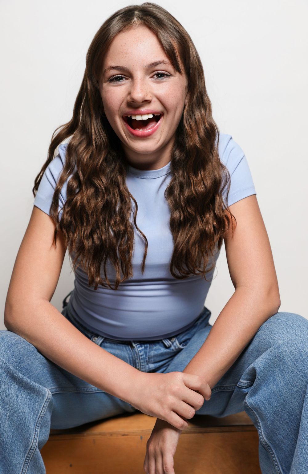 Smiling young girl with wavy brown hair wearing a light blue t-shirt and jeans, sitting cross-legged on a wooden surface.