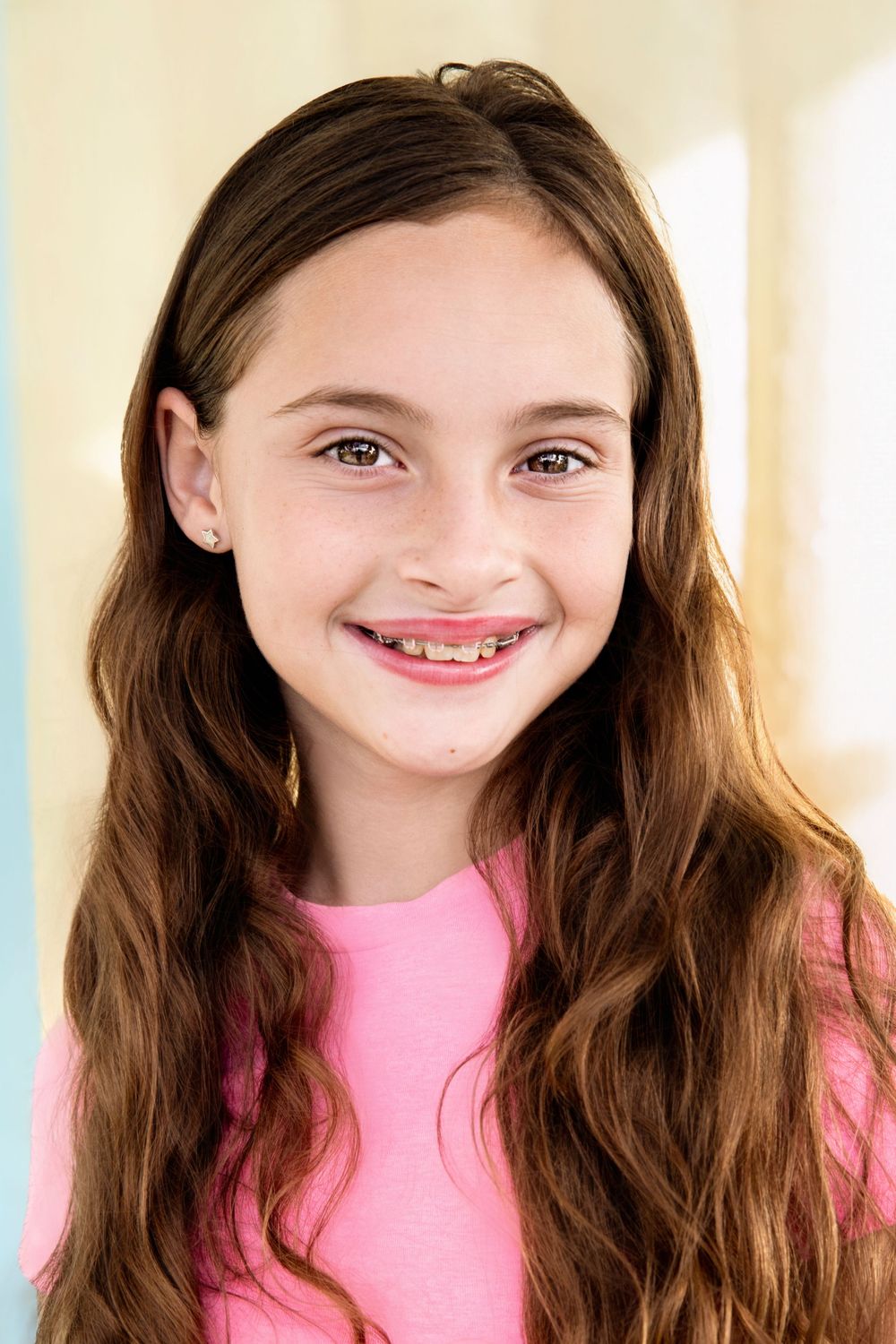Smiling young girl with long brown wavy hair wearing a pink shirt against a colorful background.