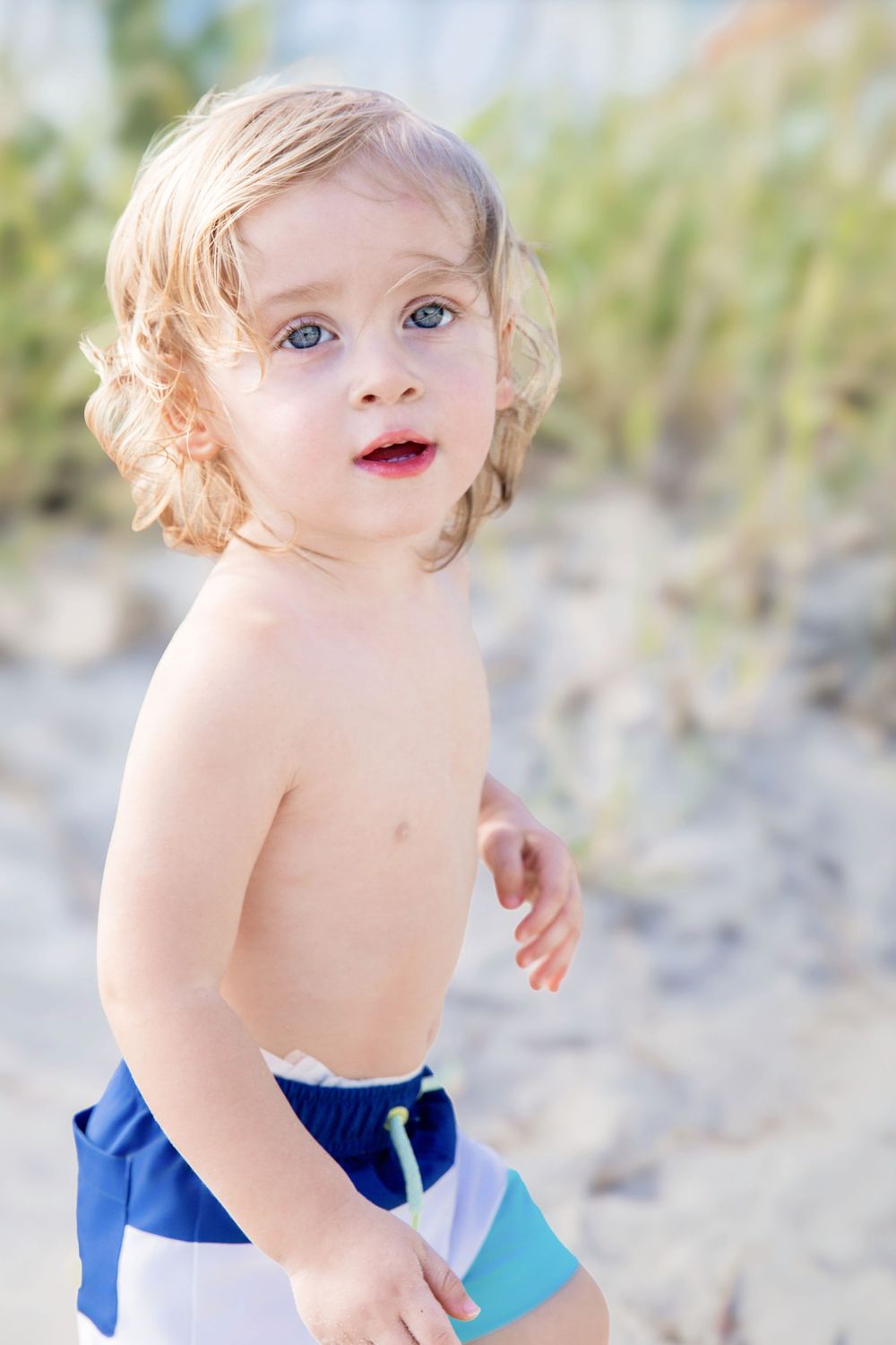 Cute toddler boy with blonde curly hair and blue eyes wearing swim trunks at the beach.