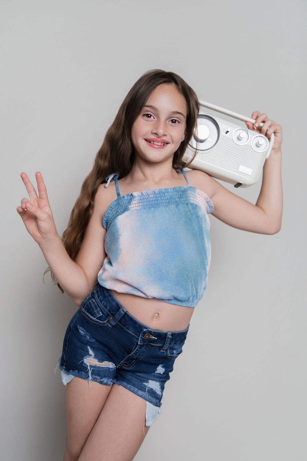 Young girl holding a retro radio boombox, making a peace sign, wearing denim shorts and a crop top.