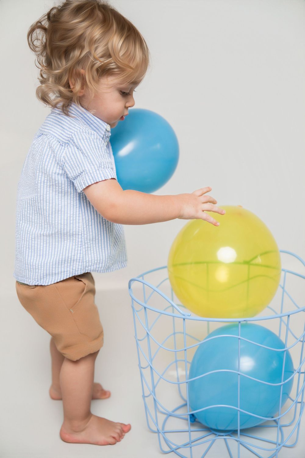 Toddler boy with curly hair playing with colorful blue and yellow balloons in a wire basket.