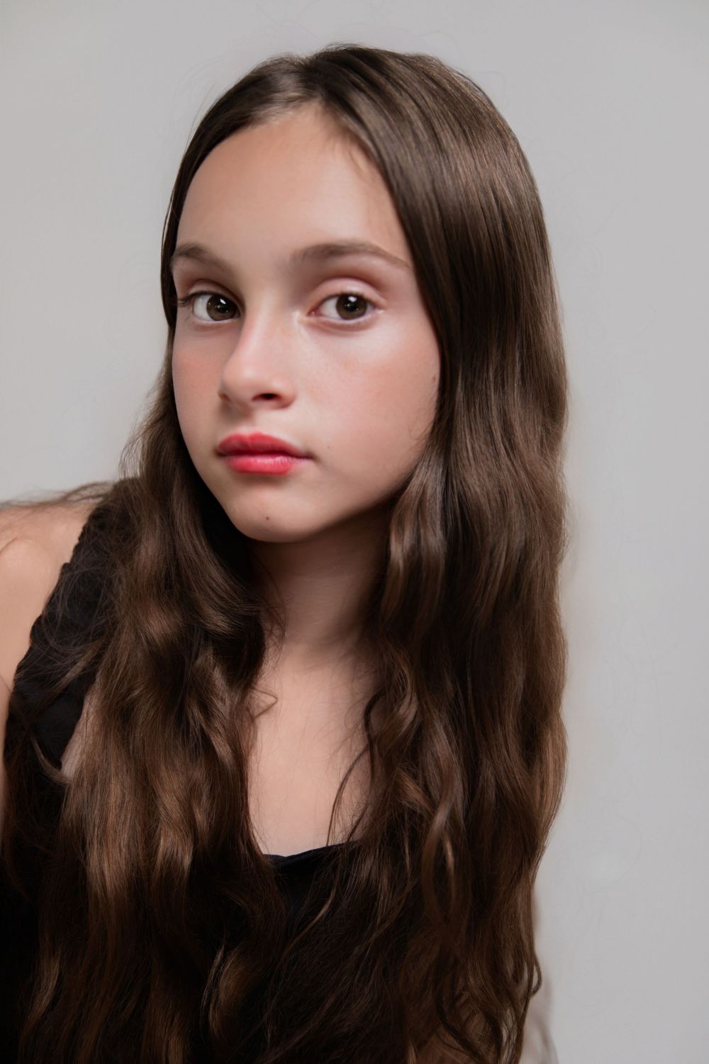 Young girl with long wavy brown hair and red lips poses against a gray background.