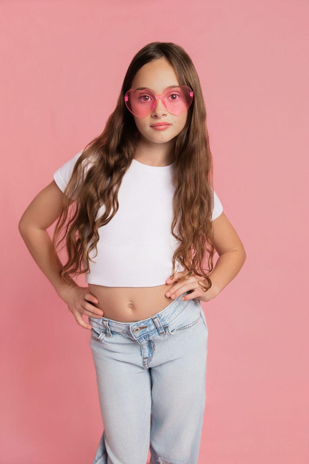 Young girl in white crop top, jeans, and pink sunglasses posing with hands on hips against a pink background.