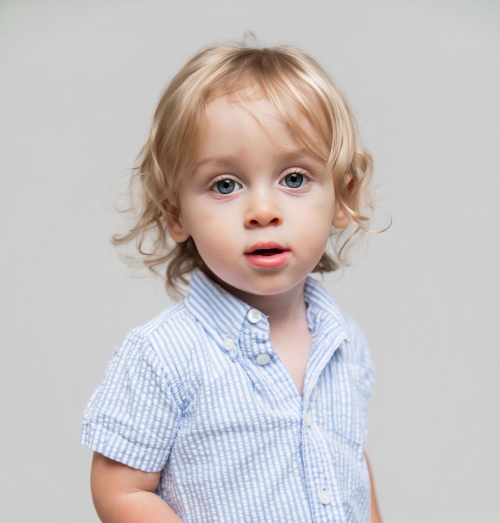 Adorable toddler with curly blonde hair and blue eyes wearing a light blue striped button-up shirt.