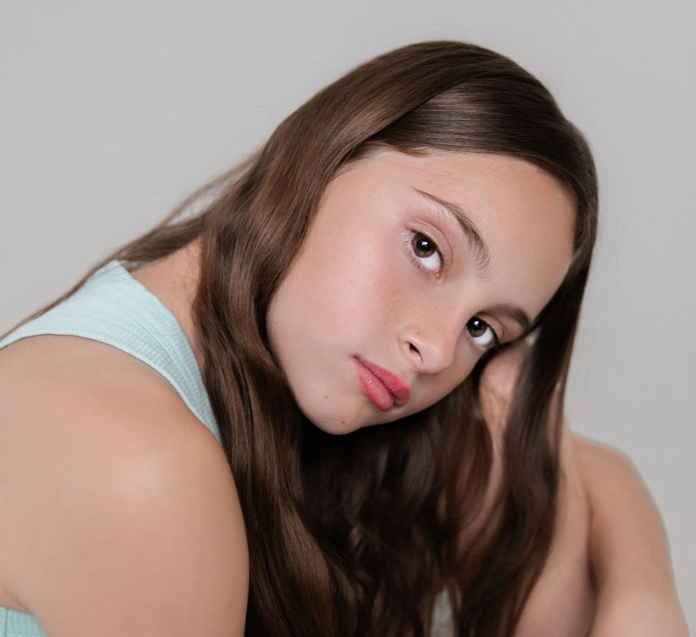Young woman with long brown hair in a light blue top, posing thoughtfully against a gray background.