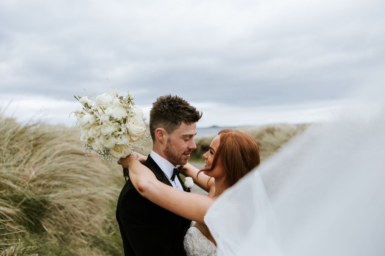 A romantic couple shares a kiss during their seaside wedding ceremony as wind sweeps through their elegant attire.