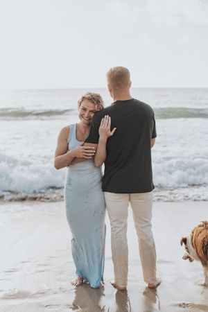 A couple shares an intimate moment by the ocean waves with their dog watching nearby.