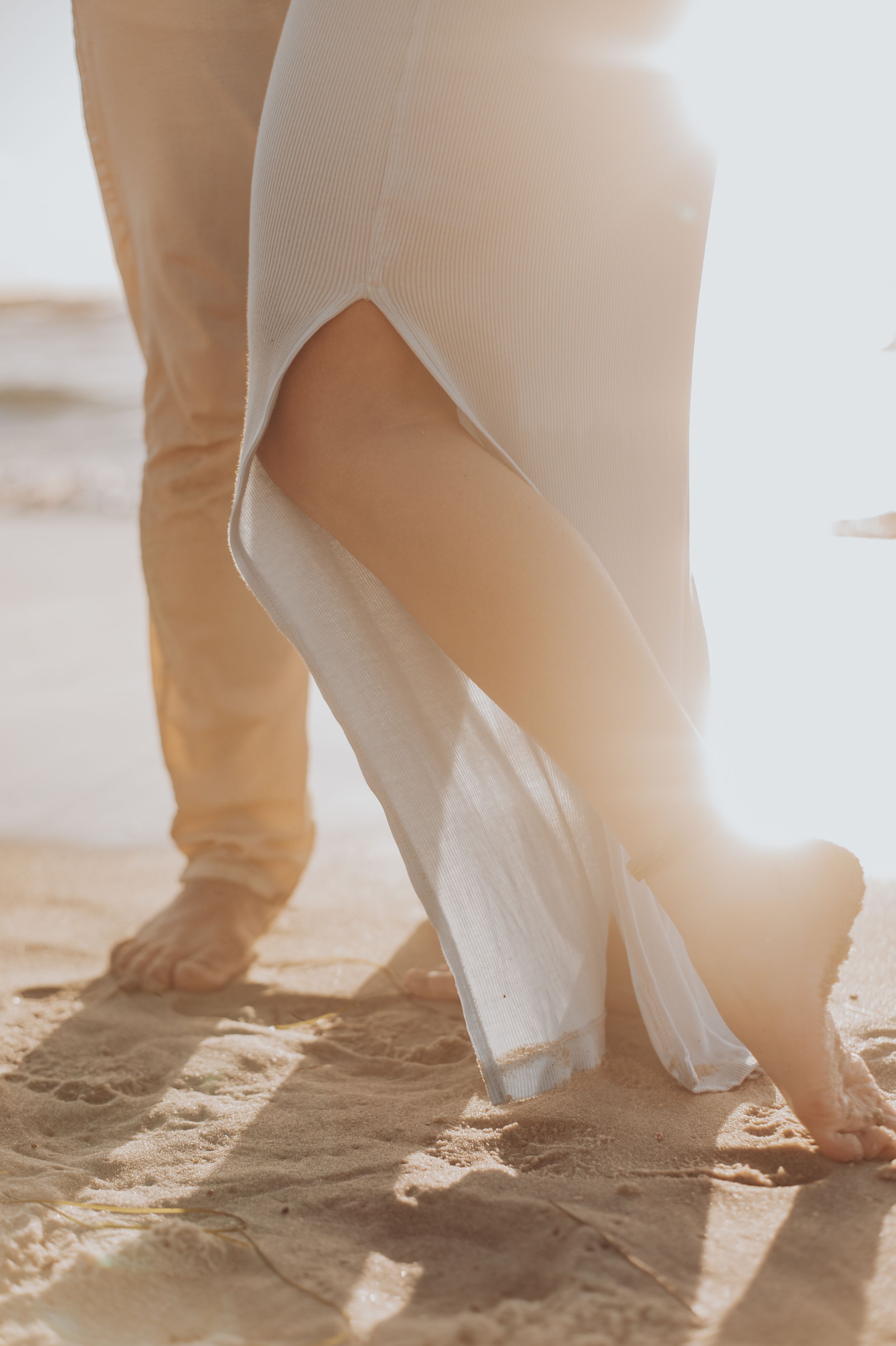 Close-up of bare feet on sandy beach with flowing white dress fabric.