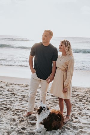 Casual beach portrait of couple in coordinated outfits with their dog on sandy shore.