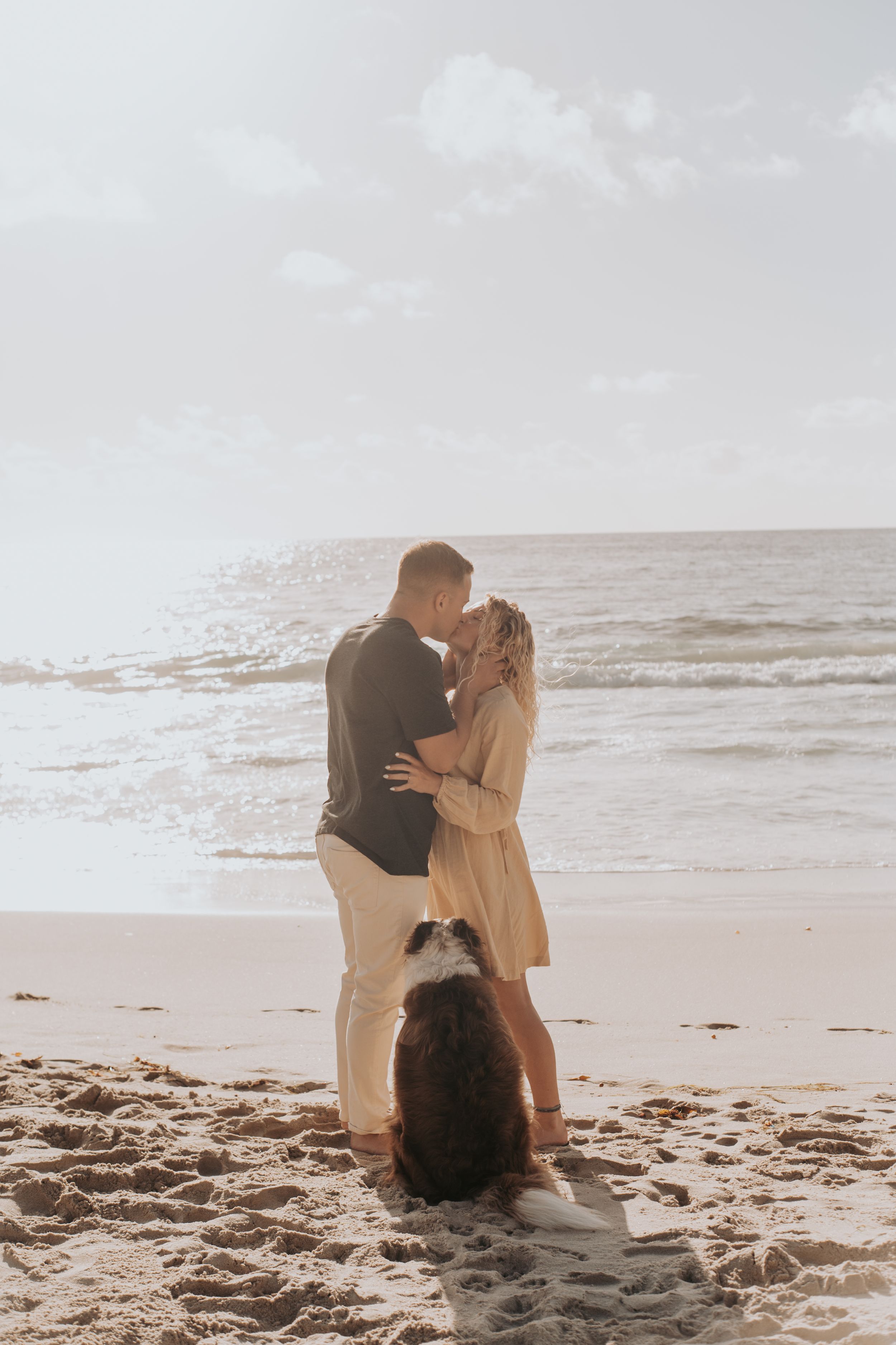 Romantic beach scene with couple embracing at sunset while dog watches nearby.