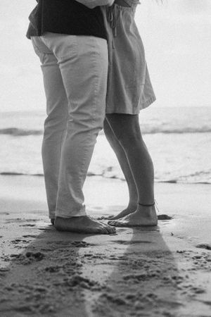 A black and white photo of two pairs of feet standing close together on wet sand.