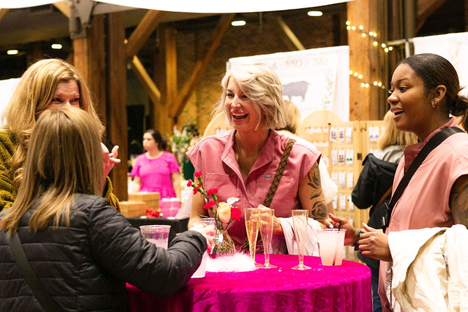 People laughing together at a social event with bright pink table decor and wooden beamed ceilings in the background.
