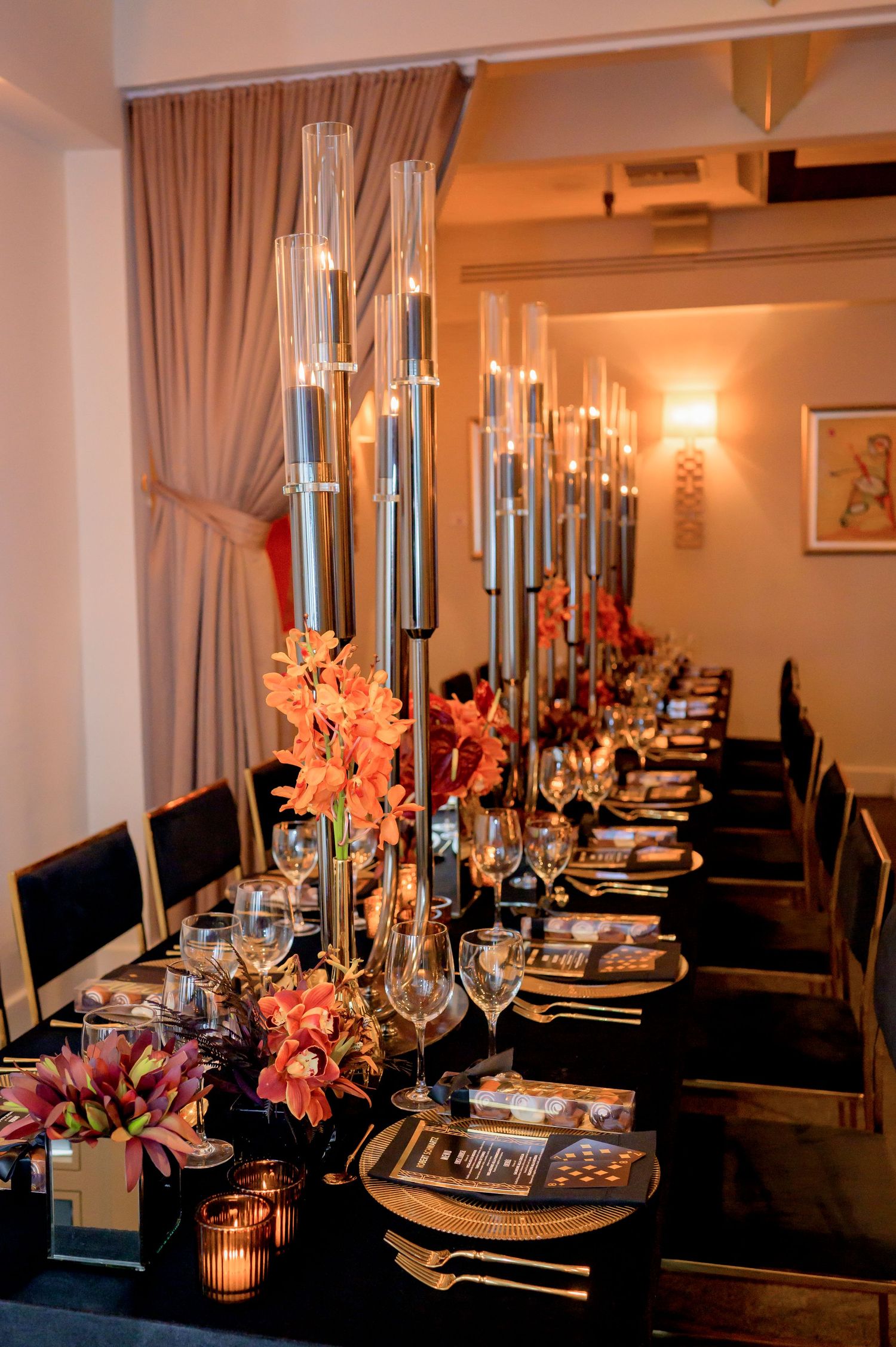 Elegant dining table with tall glass candlesticks, pink floral arrangements, and place settings on a dark tablecloth.