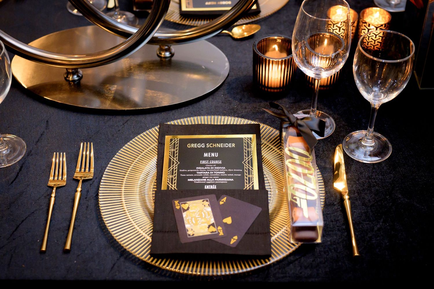 An elegant black and gold place setting with a menu card, ribbed charger plate, and gold utensils on a dark tablecloth.