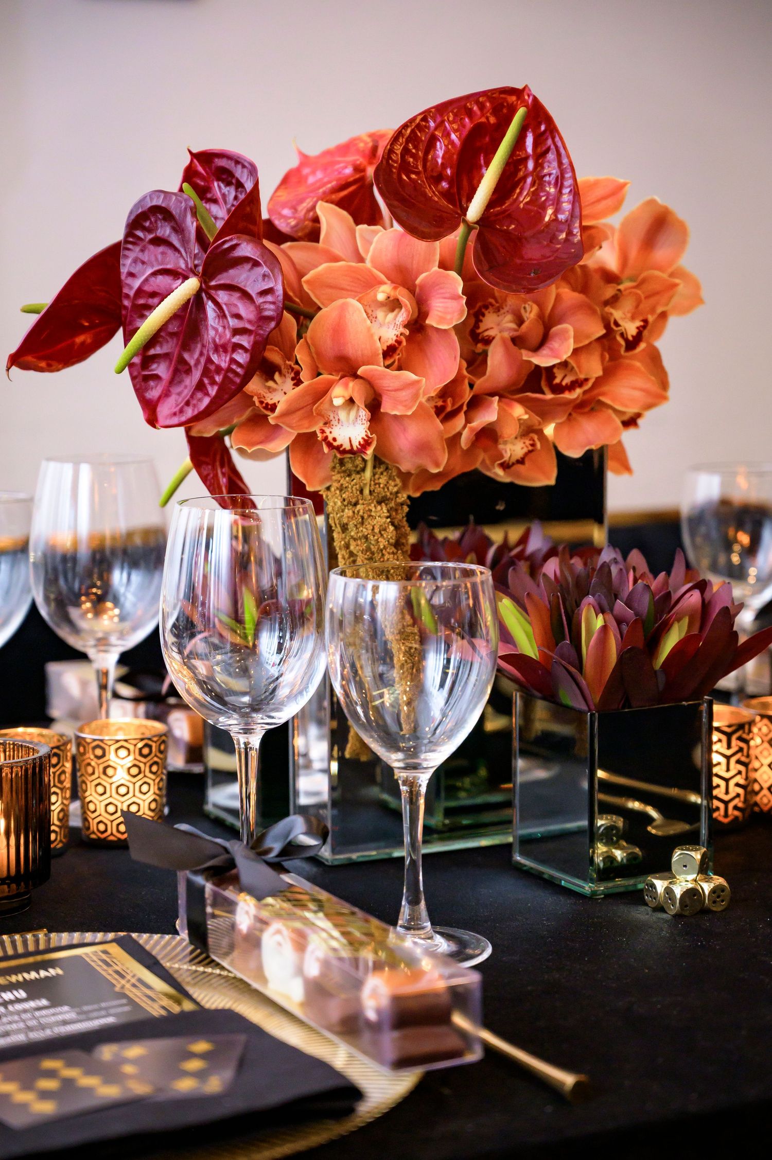 Elegant table setting with red anthurium flowers, orange orchids and wine glasses against a dark tablecloth with gold accents.