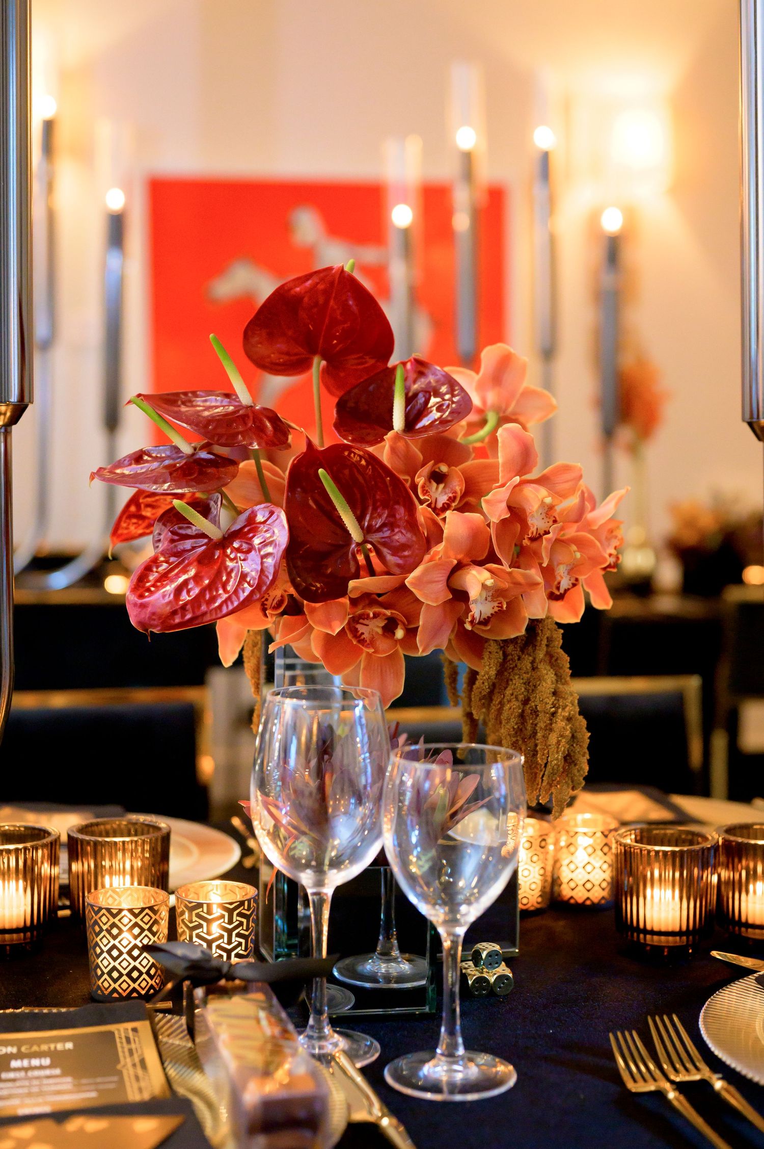 Elegant table setting with red and orange floral centerpiece, wine glasses, and glowing votive candles on dark tablecloth.