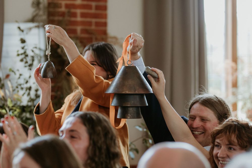 Wedding celebration scene with guests cheering and toasting in a sunlit room with exposed brick walls.
