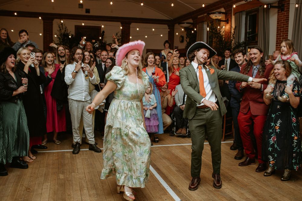 Wedding guests dance and celebrate on a wooden dance floor under twinkling lights at an indoor reception.