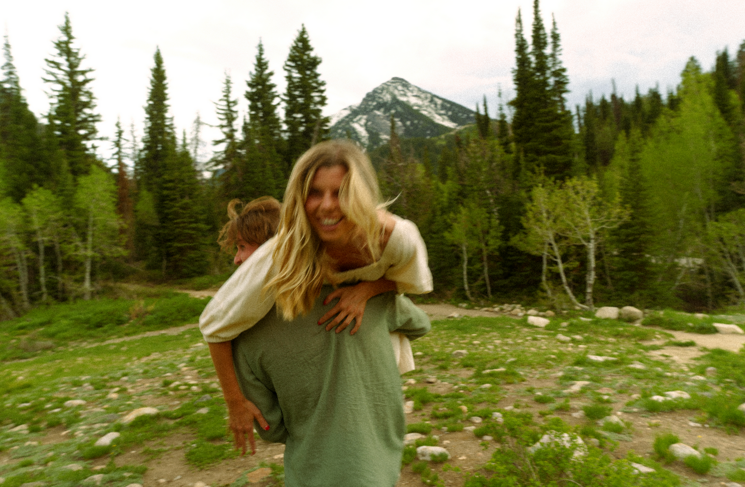 Playful piggyback ride in front of mountains and evergreen trees in a meadow.
