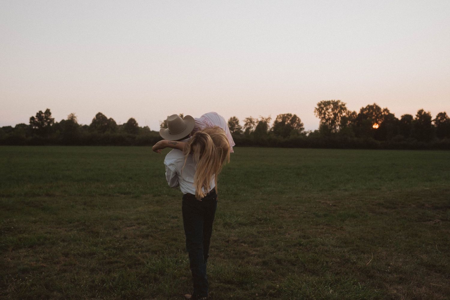 Silhouetted figure with flowing hair stands in a grassy field at sunset with trees in the distance.