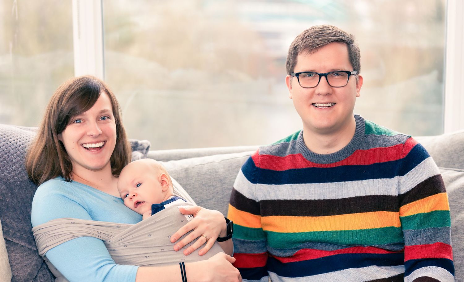 A couple sits on a couch smiling while holding a small baby in a light colored outfit.
