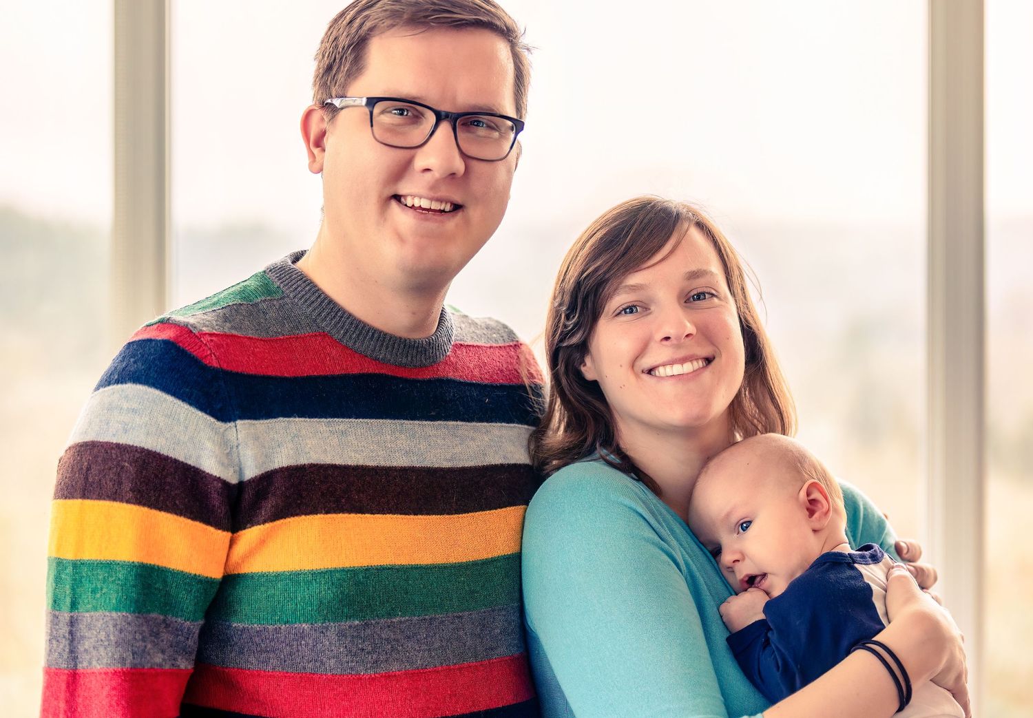 A couple in casual attire smiles while holding their sleeping baby next to bright windows.