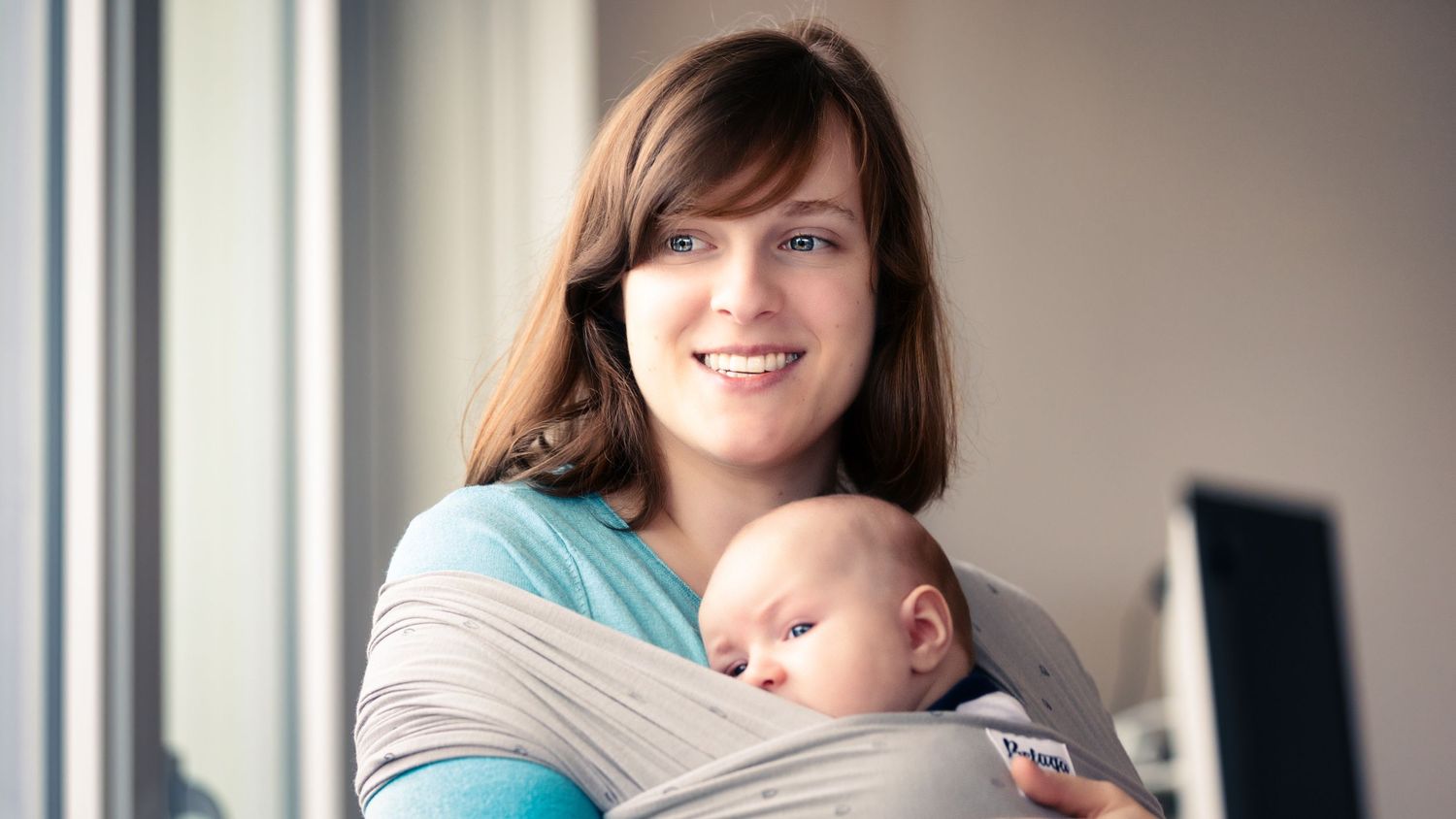 A parent wearing a light blue shirt carries a sleeping baby in a gray wrap carrier indoors.