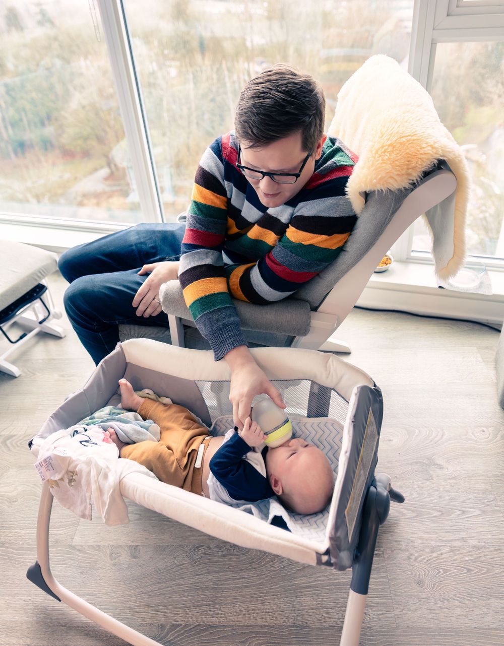 A person in a striped sweater leans over a baby resting in a bouncer seat by a sunny window.