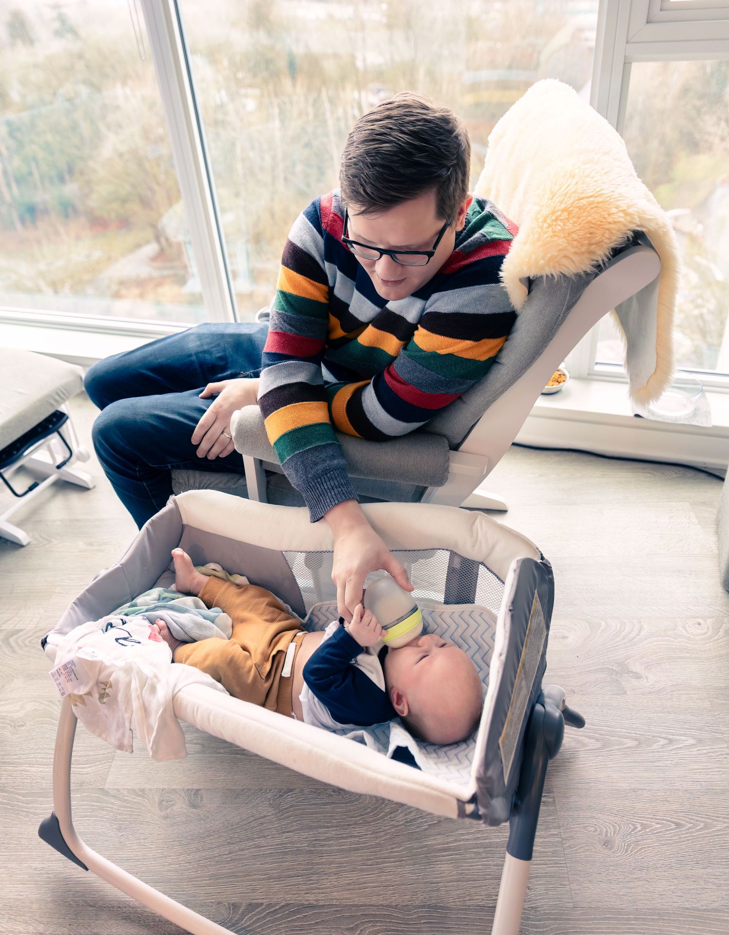 A person in a striped sweater leans over a baby resting in a bouncer seat by a sunny window.