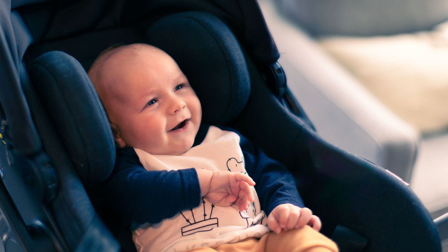 A laughing baby sits securely fastened in a navy blue car seat.
