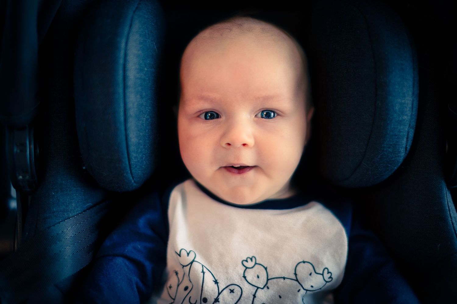 An infant wearing a white and blue bib sits secured in a dark blue car seat.