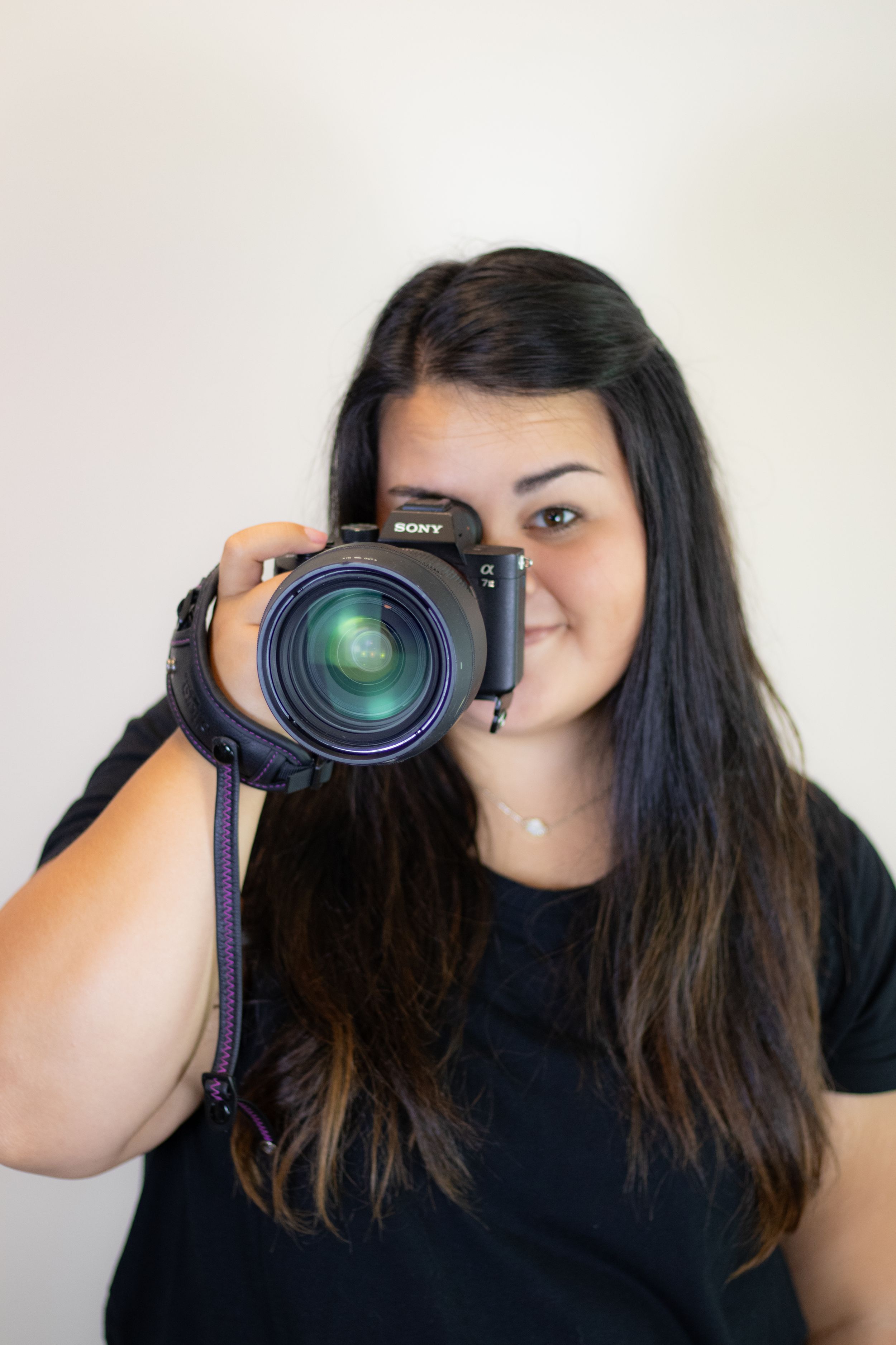 Photographer in black t-shirt holds professional Sony camera with large lens against white background.