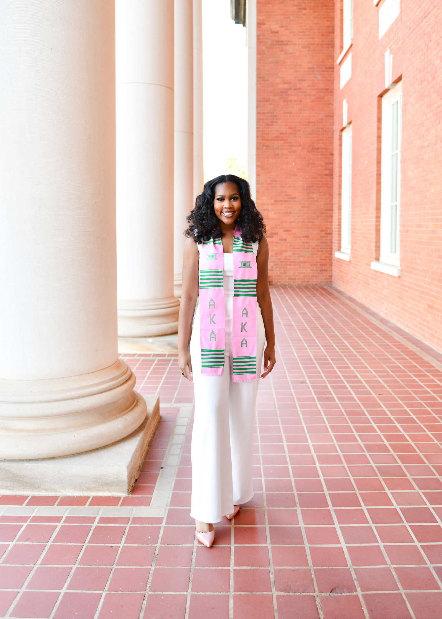Person in a pink and white patterned dress walking along a columned walkway with terracotta tiles.