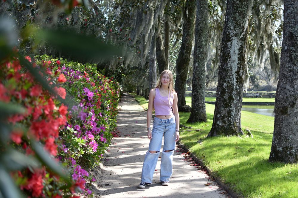 A person walks along a landscaped path surrounded by flowering bushes and moss-covered trees.