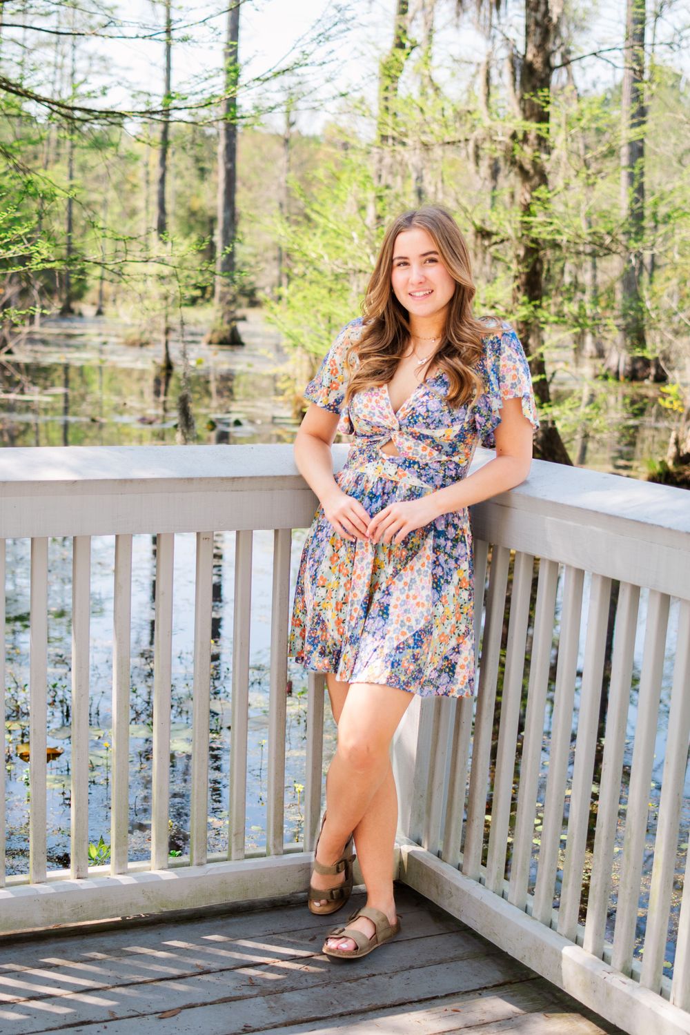 Casual portrait in floral dress leaning against white deck railing with swamp scenery.