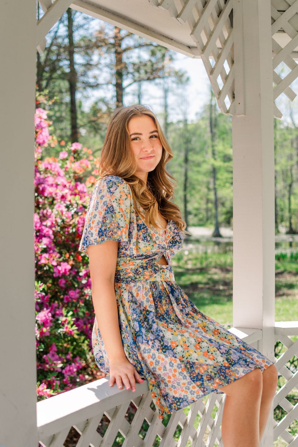A person in a floral blue dress poses on a white gazebo surrounded by pink azalea blooms and pine trees.