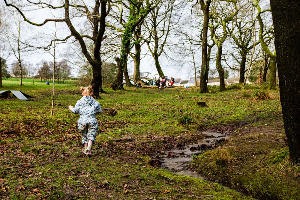 A child runs joyfully through a muddy woodland path surrounded by tall trees on a cloudy day.