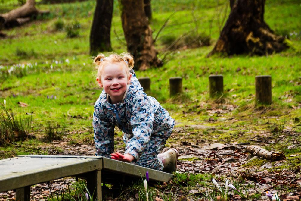 A small child in a floral jacket sits happily on a wooden bench in a forest setting.