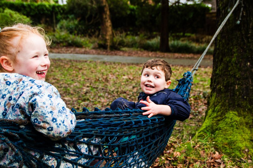 Two children laugh while swinging in a blue hammock outdoors.